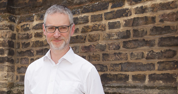 Image of man in glasses and a white button up shirt in front of a brick wall