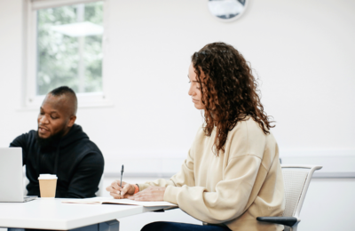 Female student is writing in a notebook and a man is sat next to them on a laptop