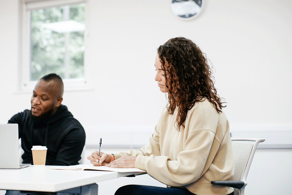 Female student is writing in a notebook and a man is sat next to them on a laptop