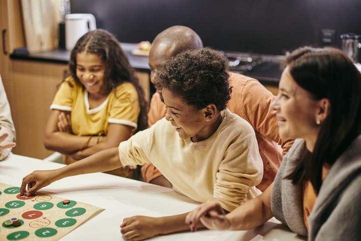 A smiling boy places a chip on a board game whilst playing it with his family. There are two adults and a young girl smiling and sat around the table with him.