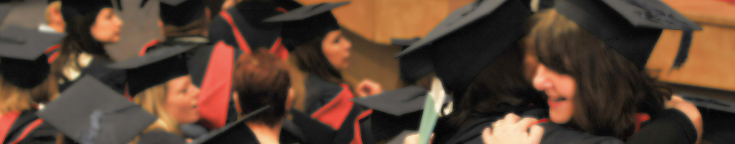Two graduands in black mortar boards and gowns embrace at graduation ceremony in a large hall